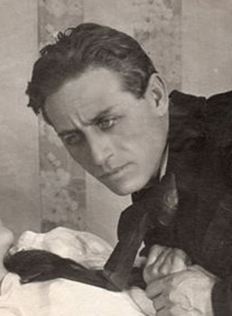 Grigori Chmara, 1918 picture. Black and white. He is holding the hand of a woman who is laying down. He is wearing a dark suit.