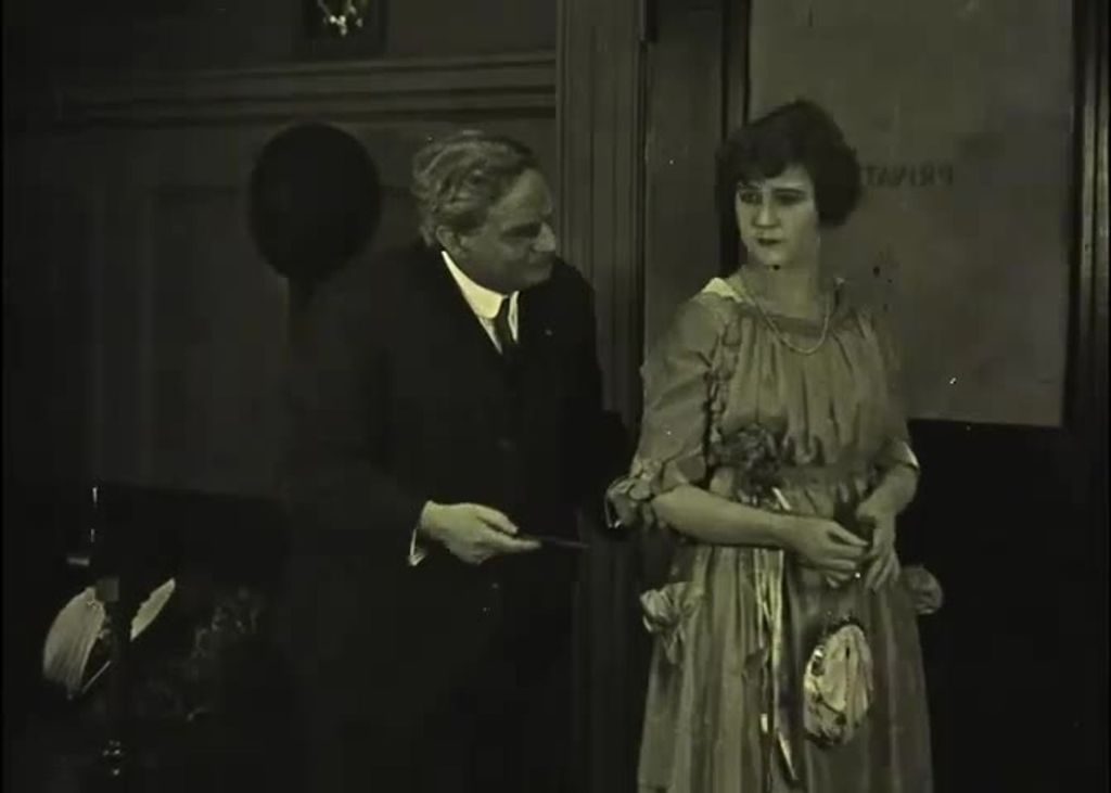 A man and a woman are in the office. He is wearing a suit, a tie and trousers. She is wearing a dress, a pearl necklace. Her hair is short and she is holding a purse.