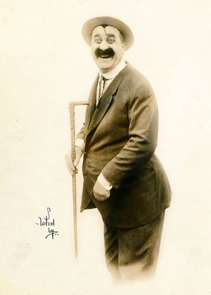 Mack Swain 1920 Promotional photo, black and white. He has a moustache. He is wearing a hat, a tie, a suit and he is holding a can. There is also his signature on the photo.