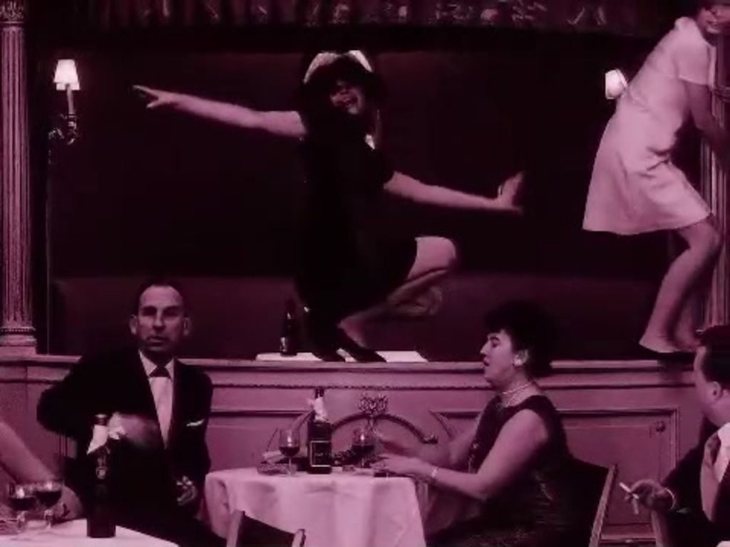 Some people are eating by a table while some women dance around them.