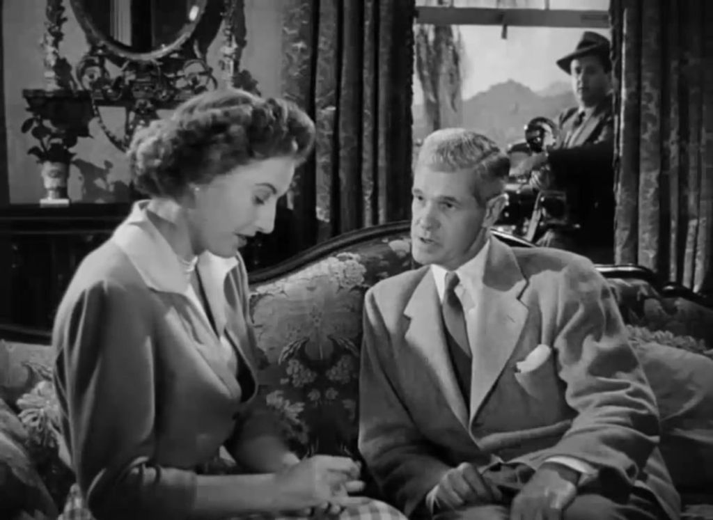 A man and a woman are sitting on a sofa. There is a man outside behind them.