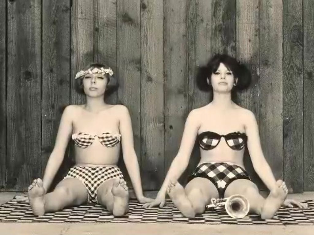 Two women are sitting down on the floor. They are wearing bikinis and their hair is short. One of women is also wearing flowers in her hair.