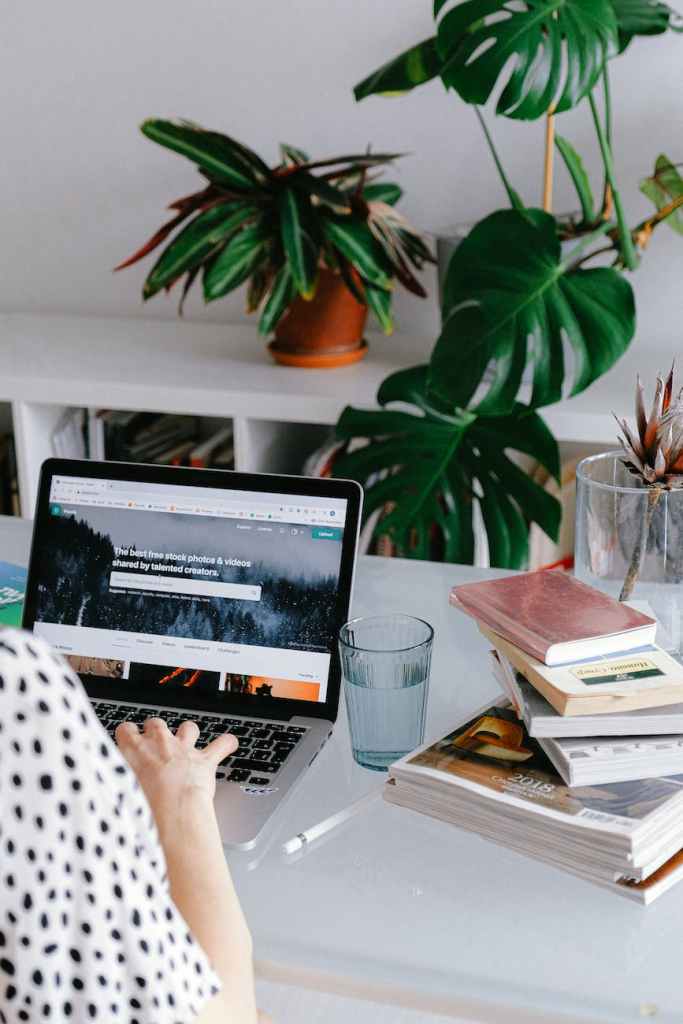 A woman is sitting by a desk. We can see a laptop, some books and a glass of water between the books and the laptop.