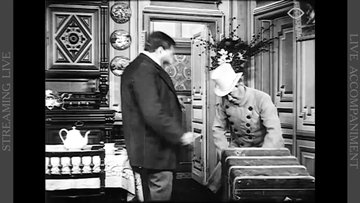 A man and a woman are facing each other. They are indoors. She is wearing a hat and long-sleeved dresses. He is wearing a suit.