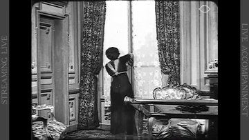 A woman is by the window of a house. We can see around her a door, a desk, a chair and curtains.