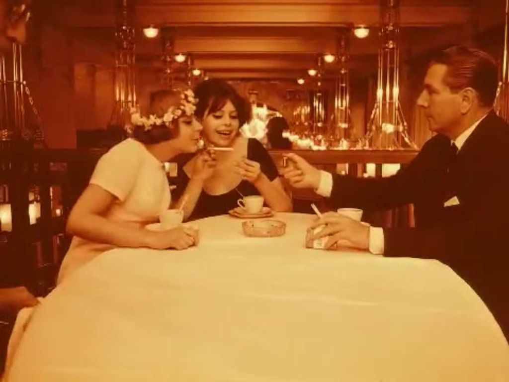 Two women and one man are in a restaurant, drinking coffee. The man is lighting the cigarette of one of the women.