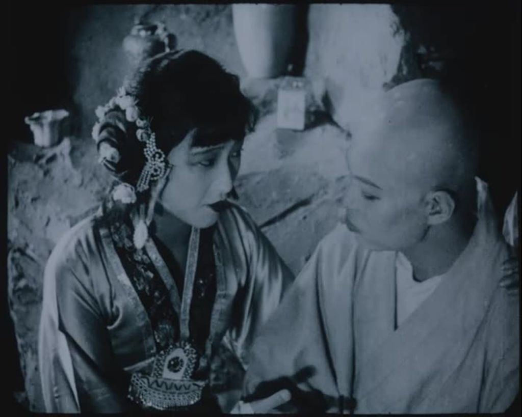 A man and a woman are wearing traditional Chinese clothes. Her hair is dark, straight, tied and there is a fringe. The man is bald and is sitting close to the woman.